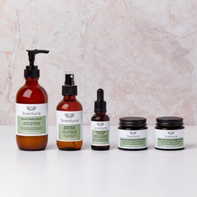 Scenturie Essential Natural Skincare Set featuring 5 award-winning amber glass bottles: Creamy Cleanser, Toner, Face Oil, Day and Night Cream on a grey background.