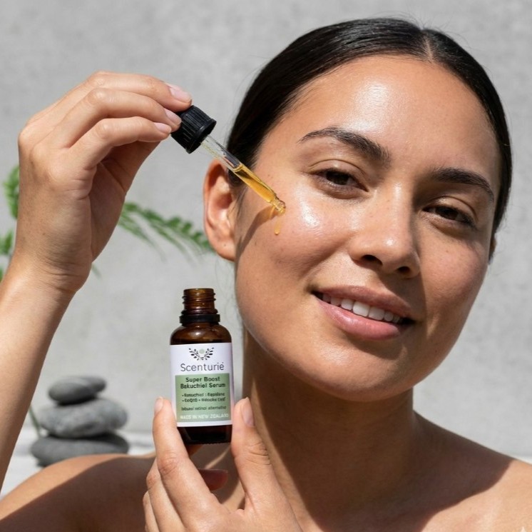 A model applying drops of Scenturie Super Boost Bakuchiol Serum to her face with a glass dropper, showcasing the CoQ10 and Kānuka Leaf formula made in New Zealand.